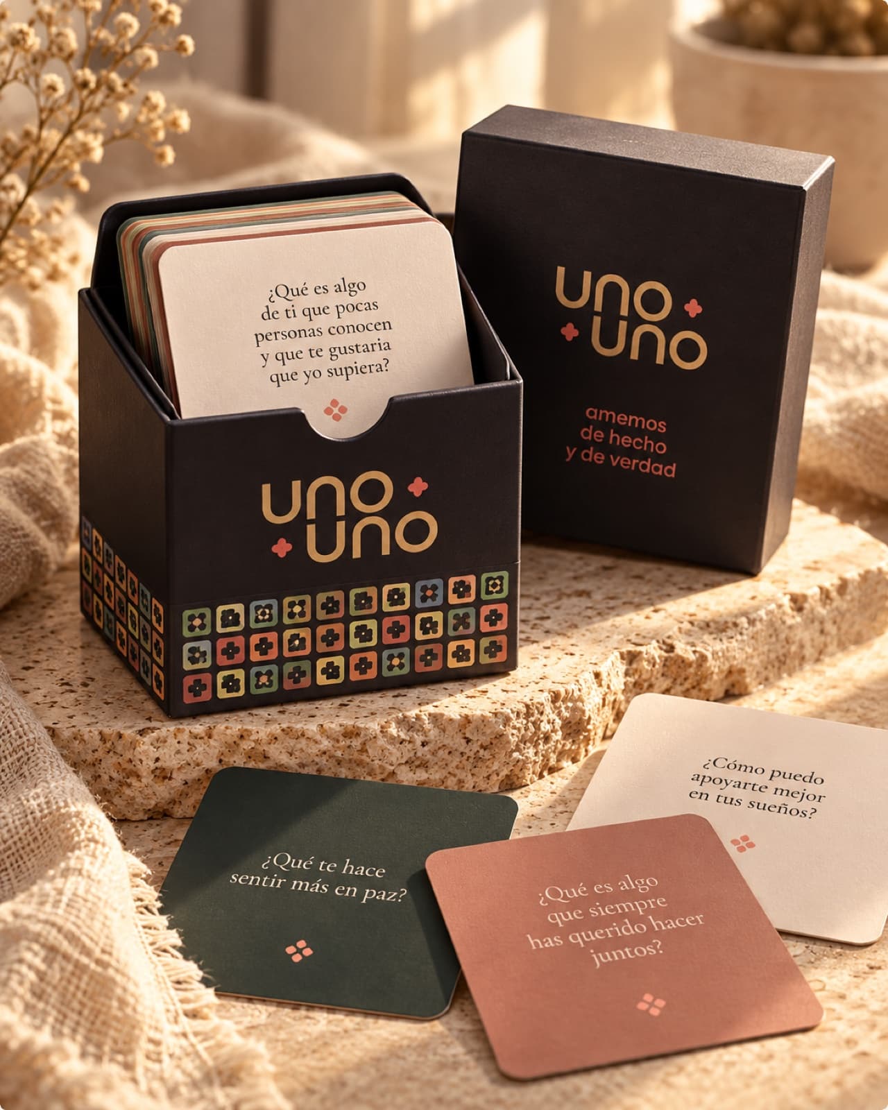 Caja de UNO+UNO con tarjetas de preguntas sobre una piedra y textiles cálidos